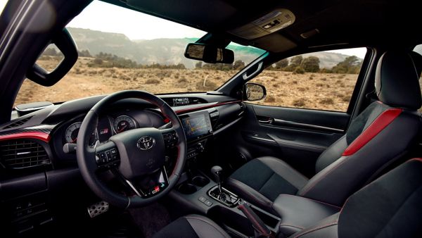 Vorderer Innenraum eines Toyota Hilux mit dunklen Sitzen und roten Akzenten, durch die Windschutzscheibe eine karge Berglandschaft.