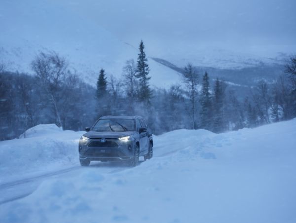 Grauer Toyota RAV4 fährt über eine mit Schnee bedeckte Straße. Es schneit und es dämmert, die Scheinwerfer des Autos sind aktiviert.